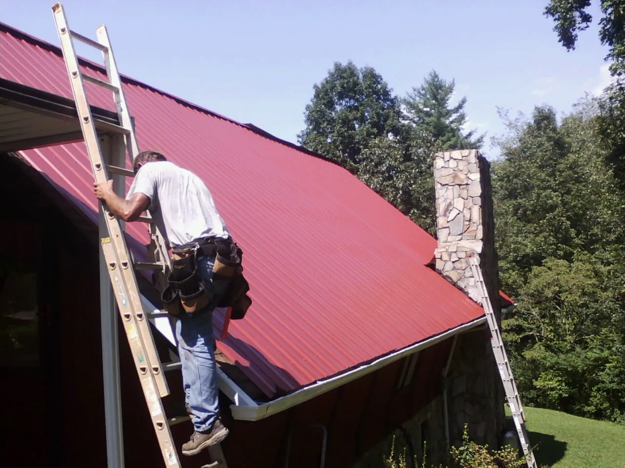 Roofer on a ladder beside a red metal roof and stone chimney — Soffit & Fascia Repair by Roofing Brothers in Nixon